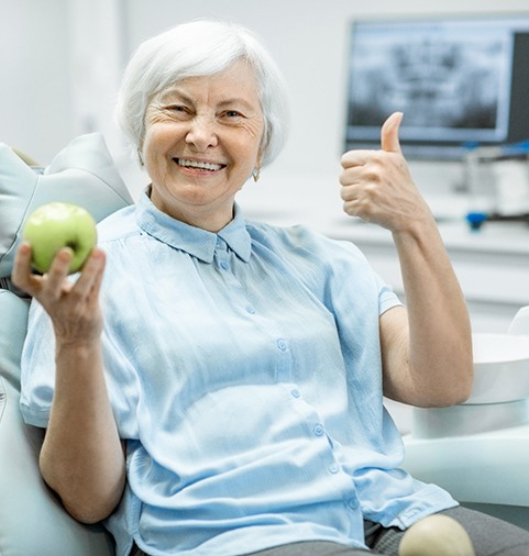 smiling woman holding an apple and giving a thumbs-up in the dental chair 